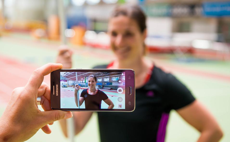 Hand hält ein Smartphone, das ein Porträt einer lächelnden Athletin mit Stab in einer Sporthalle zeigt.