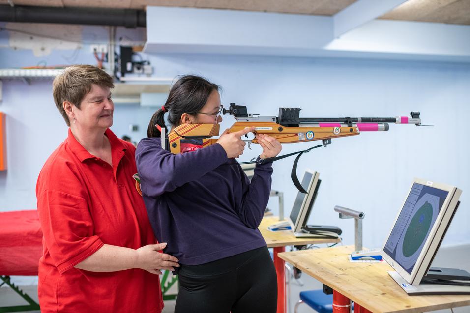 Trainerin hilft Schülerin beim Zielen mit einem Sportgewehr vor zwei Monitoren in einem Schießstand.