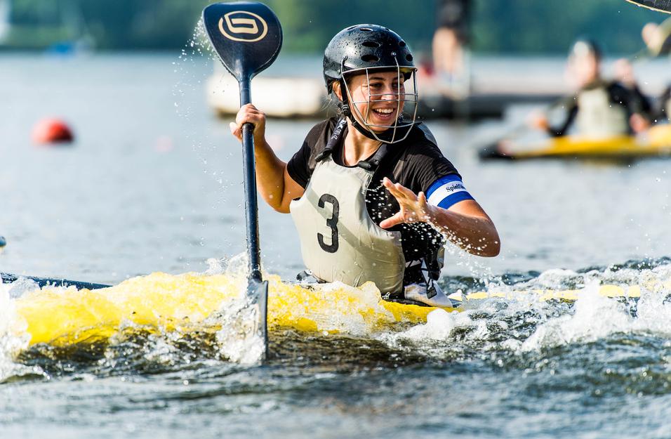 Junge Frau paddelt lächelnd in einem gelben Kajak auf einem ruhigen Gewässer, umgeben von weiteren Sportlern.